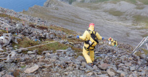 Scientists and technicians wearing safety gear including harnesses while removing PCBs along a steep slope with peaks, cliffs, and ocean in the background