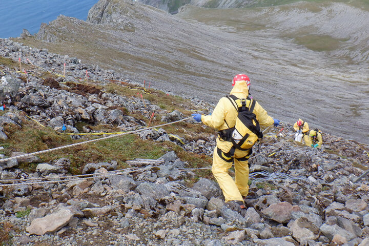 Scientists and technicians wearing safety gear including harnesses while removing PCBs along a steep slope with peaks, cliffs, and ocean in the background