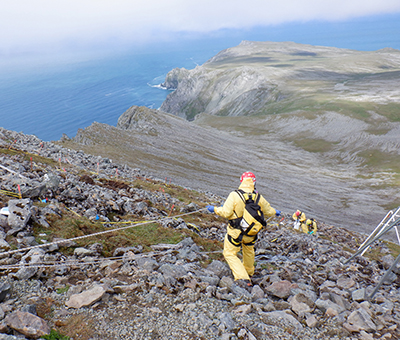 Scientists and technicians wearing safety gear including harnesses while removing PCBs along a steep slope with peaks, cliffs, and ocean in the background