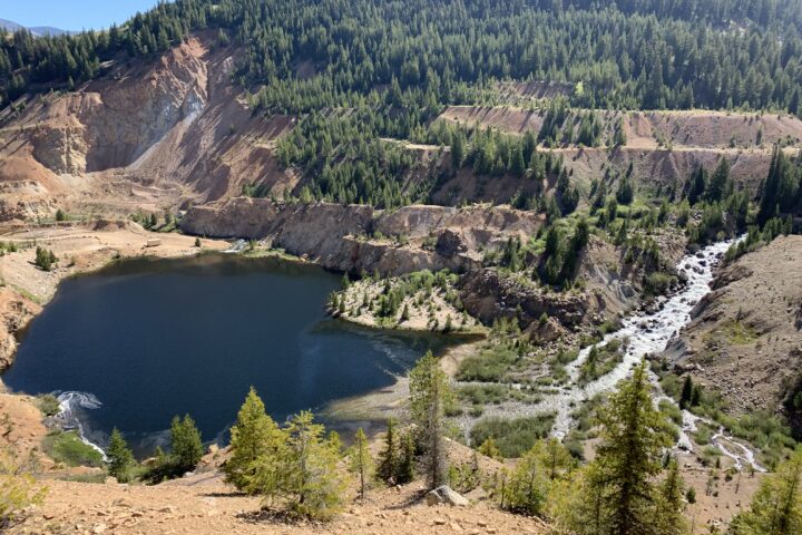 Mine remediation site shows a lake in the foreground surrounded by steep cliffs and a series of forested plateaus with a rockey river running to the lake down the right side of the screen.