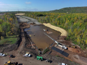 - Man-made water body utilized for subaqueous capping in the foreground surrounded by construction equipment for sediment placement with a restored floodplain and stream channel in the background.