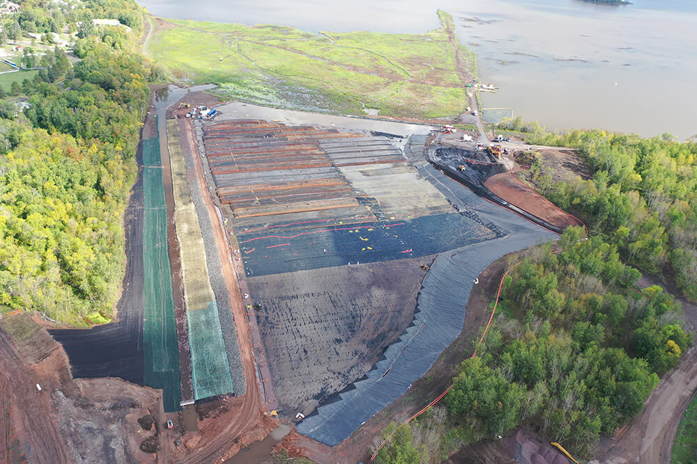 A large earthwork construction area featuring a crane and geotextile tubes used for dewatering hydraulically dredged sediment