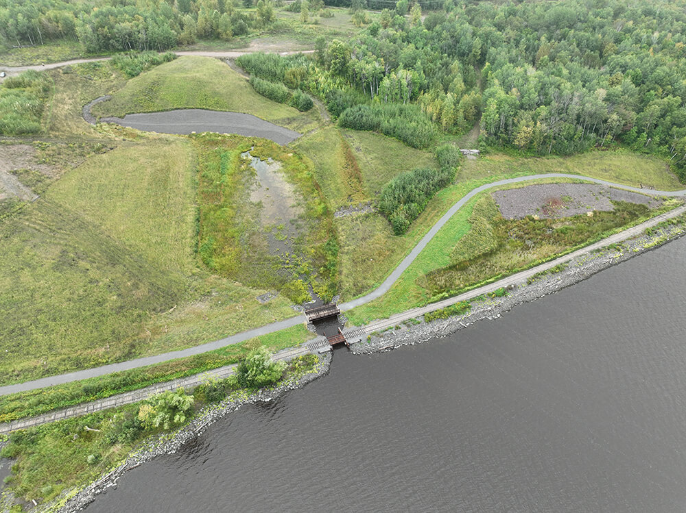 a shoreline with a railroad line next to the shoreline and a paved trail further inland followed by an oblique aerial of a thin peninsula surrounded by water.