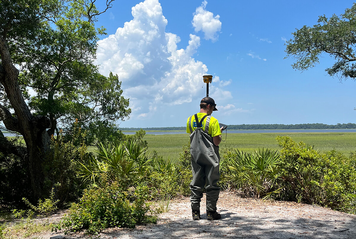 An EA scientist in waders holding a global positioning survey rod and entering data into an attached table at the edge of a marsh with open water in the distance.
