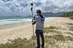 A scientist holding global positioning survey equipment on a beach in hawaii with mountains and waves in the background
