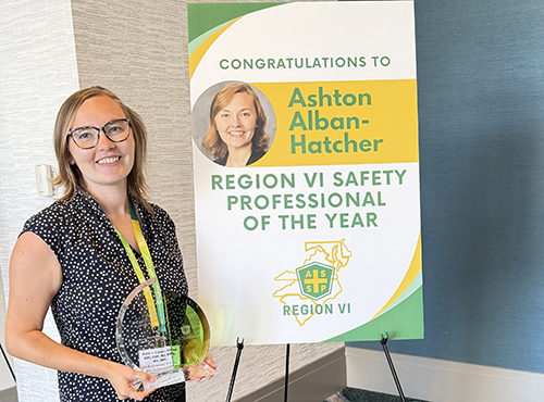 • Ashton Alban-Hatcher standing holding the Region IV Safety Professional of the Year Award standing next to a congratulatory board.